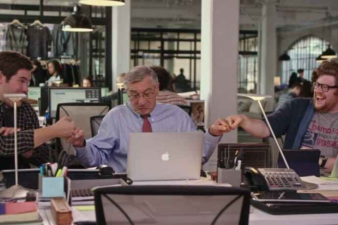 Still from the movie the Intern, showing Robert de Niro as an office intern, fist bumping the two younger men sitting on either side of him.
